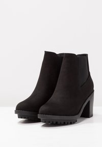 Ein Paar schwarze Wildleder-Stiefeletten mit klobigen Absätzen und elastischen Seiteneinsätzen auf einer weißen Fläche und Hintergrund.