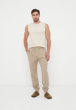 Mand iført ærmeløs beige skjorte, lysebrune joggingbukser med snøre og brune slip-on sko, stående mod hvid baggrund.