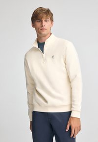 Sudadera con cremallera color crema claro, hecha de una tela suave, con cuello alto, puños acanalados y un pequeño logo bordado en el pecho.