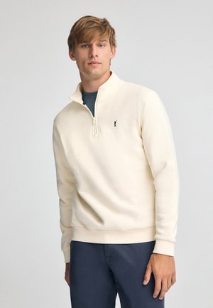 Sudadera con cremallera color crema claro, hecha de una tela suave, con cuello alto, puños acanalados y un pequeño logo bordado en el pecho.