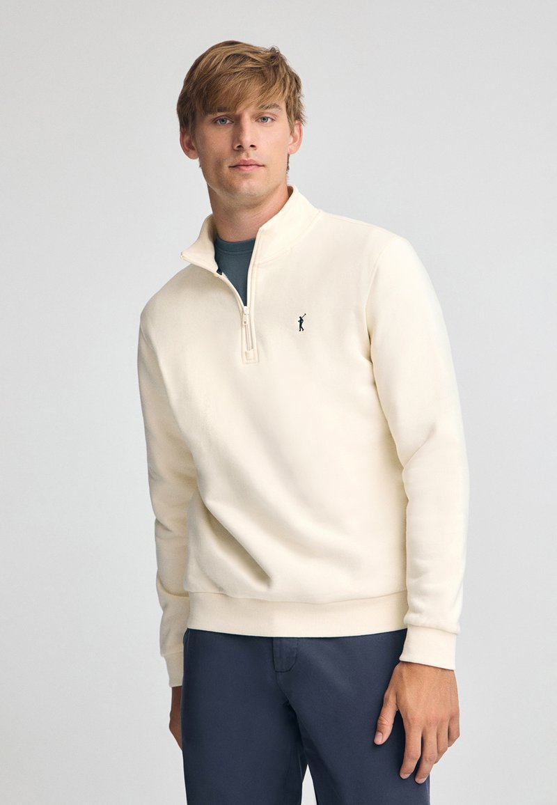 Sudadera con cremallera color crema claro, hecha de una tela suave, con cuello alto, puños acanalados y un pequeño logo bordado en el pecho.