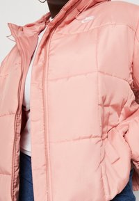 Ljust rosa Nike pufferjacka med dragkedja, kviltad struktur och fast huva. Har en liten logotyp på övre vänstra bröstområdet.