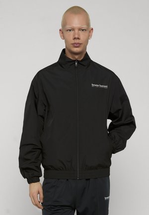 Person trägt eine schwarze Jacke mit Reißverschluss und passende Hose, beide mit "bruno banani"-Logo auf Brust und Oberschenkel, steht vor einem einfarbigen Hintergrund.