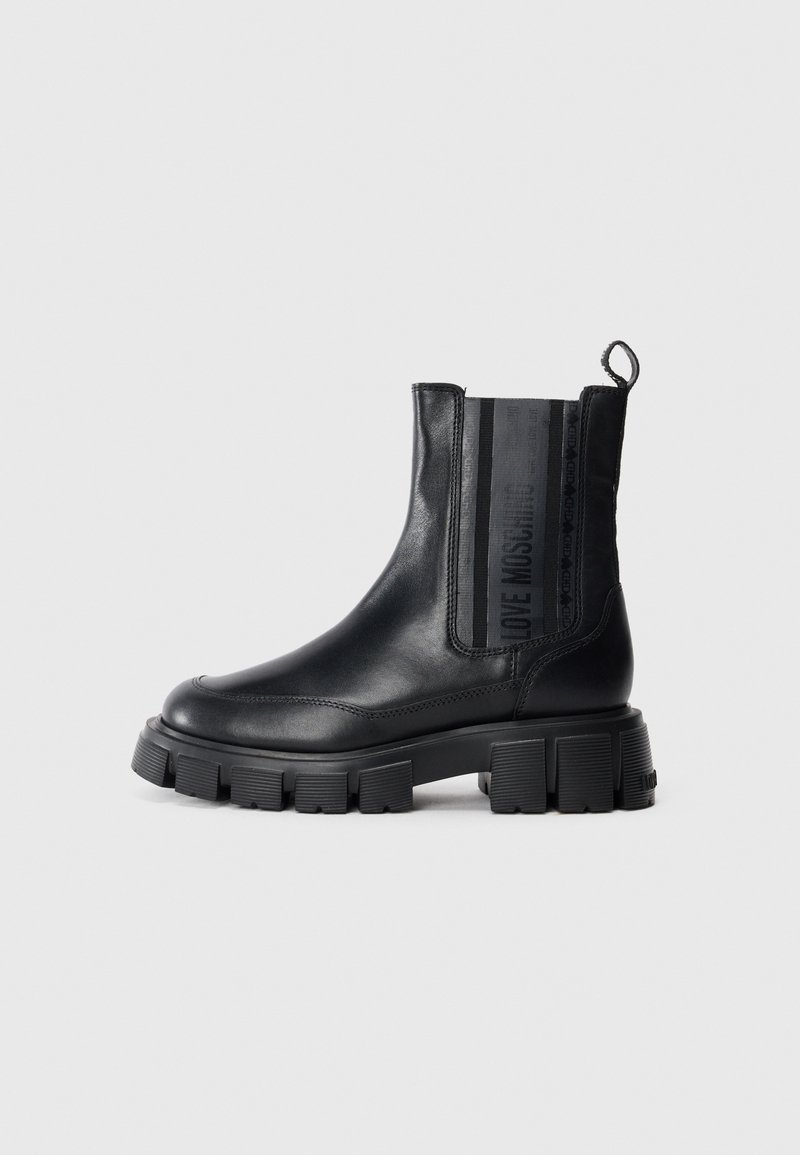Botas Chelsea de couro preto com solas de borracha robustas, apresentando painéis laterais elásticos e uma alça para facilitar o calçar. Textura suave com detalhes contrastantes.