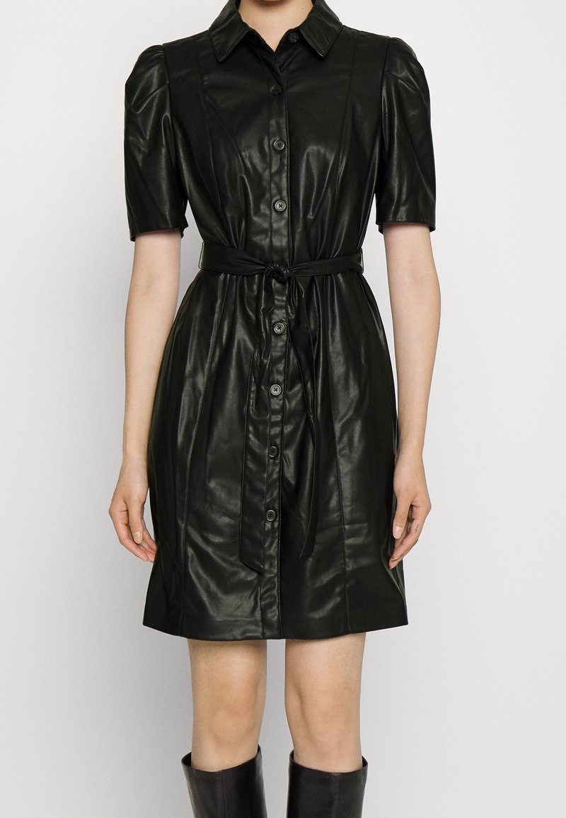 Femme portant une robe noire en simili cuir à boutons, manches courtes bouffantes, ceinture nouée à la taille et bottes noires montantes jusqu’aux genoux.