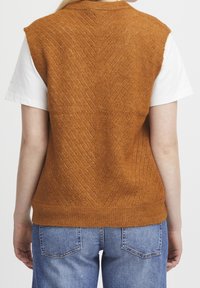 Brązowa dzianinowa kamizelka z wzorem jodełki, bez rękawów, z prążkowanym dołem, noszona na białej koszulce i połączona z niebieskimi dżinsami.