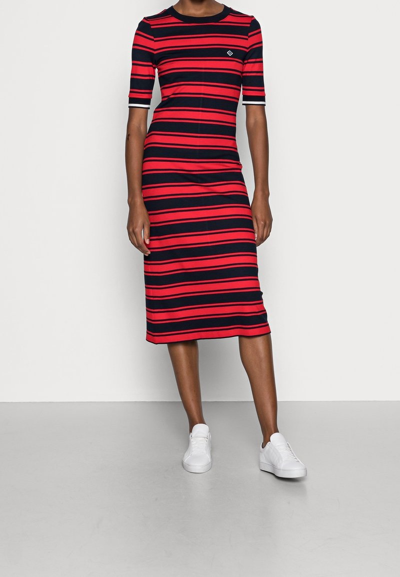Femme portant une robe mi-longue à manches courtes avec des rayures horizontales rouges et noires et des baskets blanches, debout devant un fond uni.