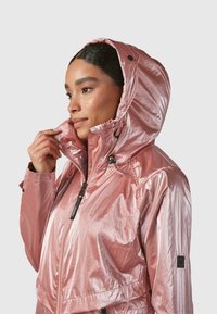 Rožinė metalinė striukė su kapišonu, užtrauktuku priekyje, reguliuojamu apykakle ir logotipo pažymėjimu ant rankovės. Lygus, blizgus paviršius.