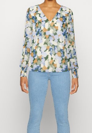 Femme portant un chemisier à manches longues et col en V avec un imprimé floral bleu, vert, blanc et beige, assorti à un jean slim bleu clair.