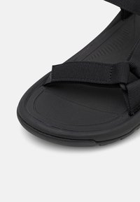 Lähikuva mustasta sandaalist, jossa on teksturoidut pohjatyynyt ja säädettävä hihna varpaiden ylle valkoisella taustalla.