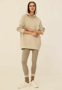 Przesadnie duża beżowa bluza z kapturem z grafiką z przodu, w połączeniu z jasnobrązowymi legginsami i białymi sneakersami. Tekstura wydaje się miękka i dziana.