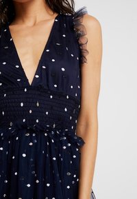 Laivastonsininen mekko syvällä V-kaula-aukolla, läpikuultavat volangihihat ja satunnaiset moniväriset pilkut textured-kankaalla. Ruttunyöritys vyötäröllä.