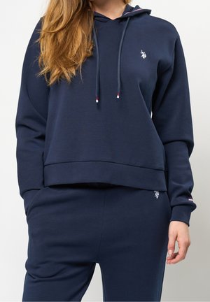 Nainen pukeutunut tummansiniseen huppariin ja siihen sopiviin verkkareihin, joissa on pieni valkoinen kirjailtu logo rinnassa ja reidessä.