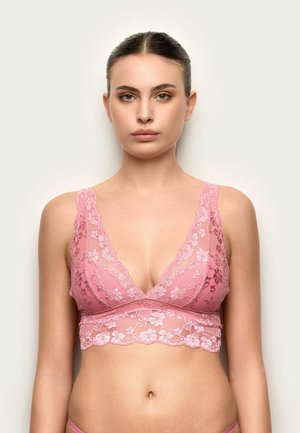 Junge Frau mit zurückgebundenen Haaren trägt einen rosa floralen Spitzen-Bralette und steht vor einem schlichten hellen Hintergrund, nach vorne blickend.