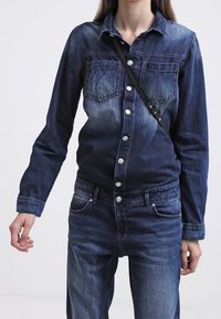 Persona que lleva una chaqueta de mezclilla oscura ajustada con bolsillos frontales y jeans a juego, llevando una bandolera negra.