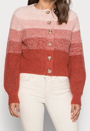 Femme portant un cardigan en tricot côtelé boutonné avec un dégradé de couleur du rose au rouge rouille et un pantalon blanc taille haute.