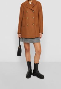 Bruine dubbelzijdige jas met gouden knopen, twee zijzakken, gedragen over een geruite rok. Zwarte enkellaarzen en een kleine zwarte handtas maken de look compleet.