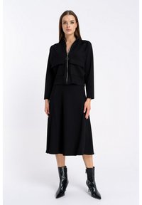 Czarna krótka kurtka z zamkiem błyskawicznym z przodu, dwoma dużymi naszytymi kieszeniami, w połączeniu z czarną spódnicą do kolan i spiczastymi czarnymi botkami za kostkę.