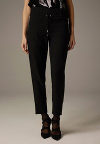 Schwarze, maßgeschneiderte Hose mit elastischem Bund, seitlichen Reißverschlusstaschen und schmal zulaufendem Bein. Weicher Stoff mit glatter Textur und ohne sichtbare Muster.