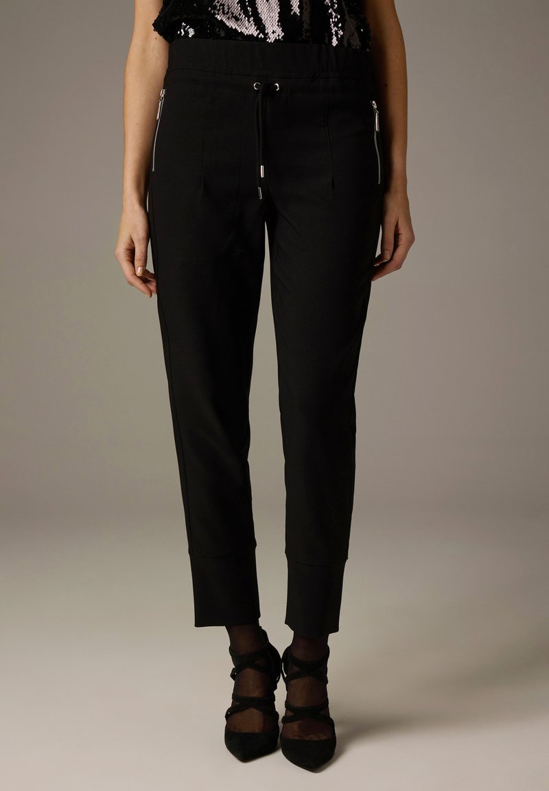 Schwarze, maßgeschneiderte Hose mit elastischem Bund, seitlichen Reißverschlusstaschen und schmal zulaufendem Bein. Weicher Stoff mit glatter Textur und ohne sichtbare Muster.