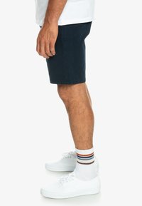 Shorts en coton bleu marine à coupe droite, portés au-dessus du genou, assortis à des baskets blanches et des chaussettes rayées.