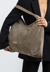 Grote, zachte suède handtas in gedempt bruin, met een brede schouderband, gebogen bovenkant en een goudkleurig logo plaatje aan de voorkant.