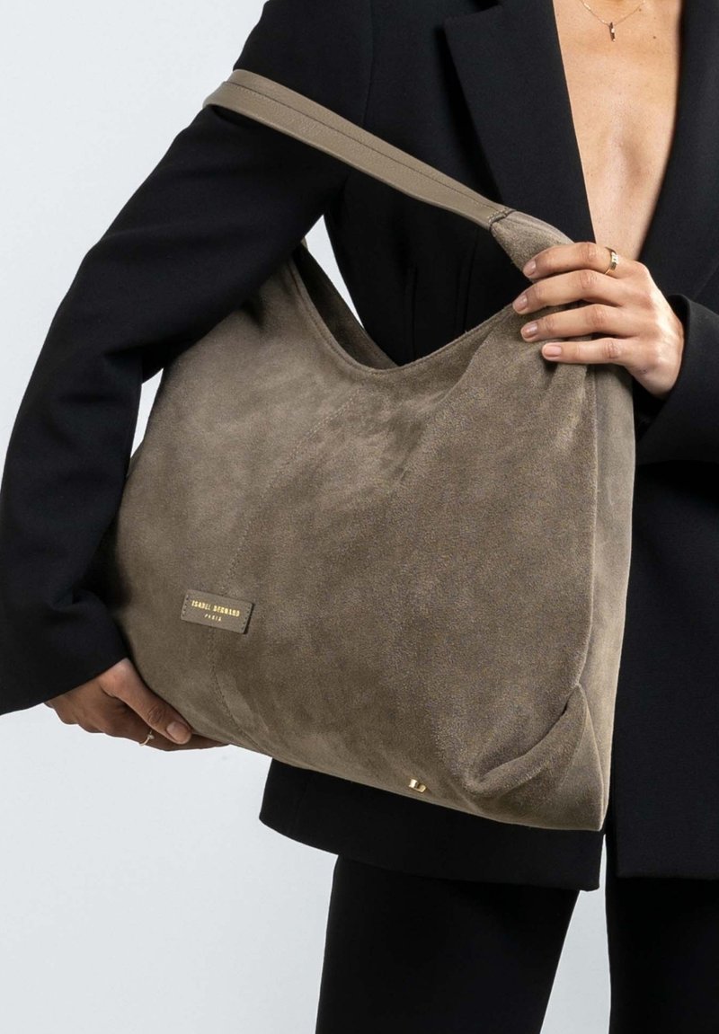 Grote, zachte suède handtas in gedempt bruin, met een brede schouderband, gebogen bovenkant en een goudkleurig logo plaatje aan de voorkant.