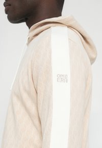 Lahka breskvasta jopa s geometrijskim vzorcem, belo poudarjeno črto na rami in izvezenim napisom "OPEN ERA" blizu izreza na vratu.