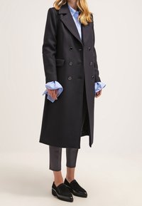 Abrigo de mezcla de lana azul marino, de corte cruzado, con solapas en pico, bolsillos laterales y puños de camisa azules en contraste. Lleva puesto sobre pantalones ajustados grises y mocasines negros.