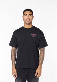 Muž s tetovanými pažemi, který má na sobě černé tričko s krátkými rukávy a červeným a bílým nápisem na hrudi, stojí před bílým pozadím.