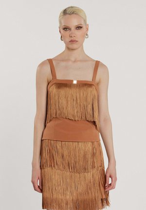 Donna con capelli biondi che indossa un vestito senza maniche color sabbia con dettagli a frange stratificate su uno sfondo semplice.