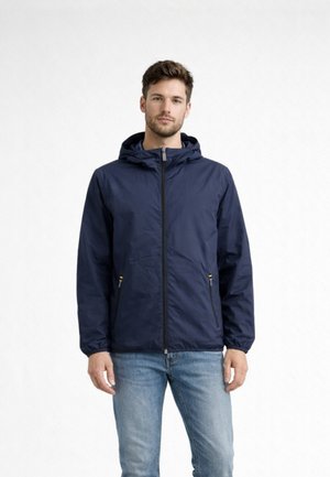 Uomo con giacca blu navy con cappuccio e tasche con cerniera, in piedi davanti a uno sfondo chiaro uniforme, con jeans azzurri.