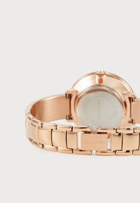 Rožinio aukso metalinis laikrodis su tekstūruota grandinėle ir apvaliu ciferblatu. Yra karūna ir išgraviruotas "Swarovski" logotipas ant sagties.