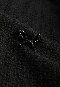 Ornement en forme de nœud orné de perles noires cousu sur un tissu noir texturé avec un motif tissé.