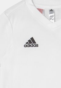 Fehér Adidas sportpóló fekete Adidas logóval, amely a V-nyakú gallér alatt középen található, és egy látható méretcímke a gallér belsejében.