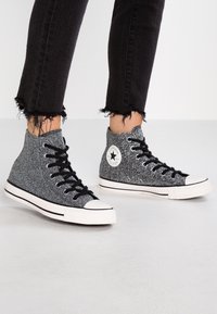Schwarze und silberne Glitzer-Hochtop-Sneaker mit schwarzen Schnürsenkeln, weißer Gummikappe und Logopatch an der Seite. Ausgefrottete schwarze Jeans.