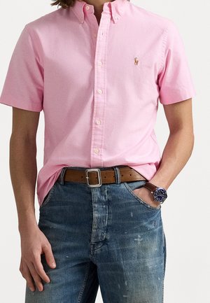 Mężczyzna ubrany w różową koszulę na krótki rękaw zapinaną na guziki, niebieskie dżinsy z brązowym paskiem oraz niebieski zegarek na rękę z brązowym skórzanym paskiem.