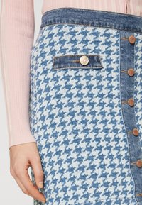 Denimskjørt med et blått og hvitt houndstooth-mønster, som har en front knappelukking, en enkel lomme og en detaljer med gullfarget knapp.
