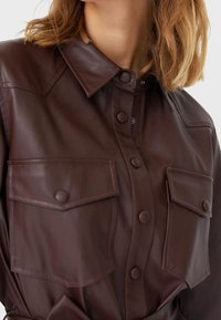 Brązowa skórzana koszula z kołnierzykiem, dużymi guzikami, dwoma przednimi kieszeniami i gładką fakturą.