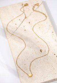 Goldkette mit kleinem Herzanhänger, feinen Gliedern, glatter Oberfläche und einem Karabinerverschluss, auf einer strukturierten Steinoberfläche präsentiert.