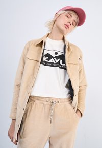 Beige Cordjacke mit Knopfverschluss und zwei Brusttaschen, getragen über einem weißen KAVU-Graphic-T-Shirt und passenden beigen Hosen mit Kordelzug.