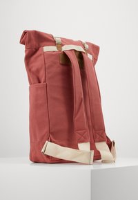 Roter Canvas-Rucksack mit beigen Trägern, Rolltop-Design, seitlicher Tasche und Lederakzenten. Verfügt über verstellbare Schulterriemen.