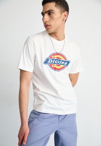 Valkoinen puuvillapaita, jossa on edessä pyöreä sininen ja punainen Dickies-logo. Mallilla on päällään laventelinväriset housut ja hopeaketju kaulassa.