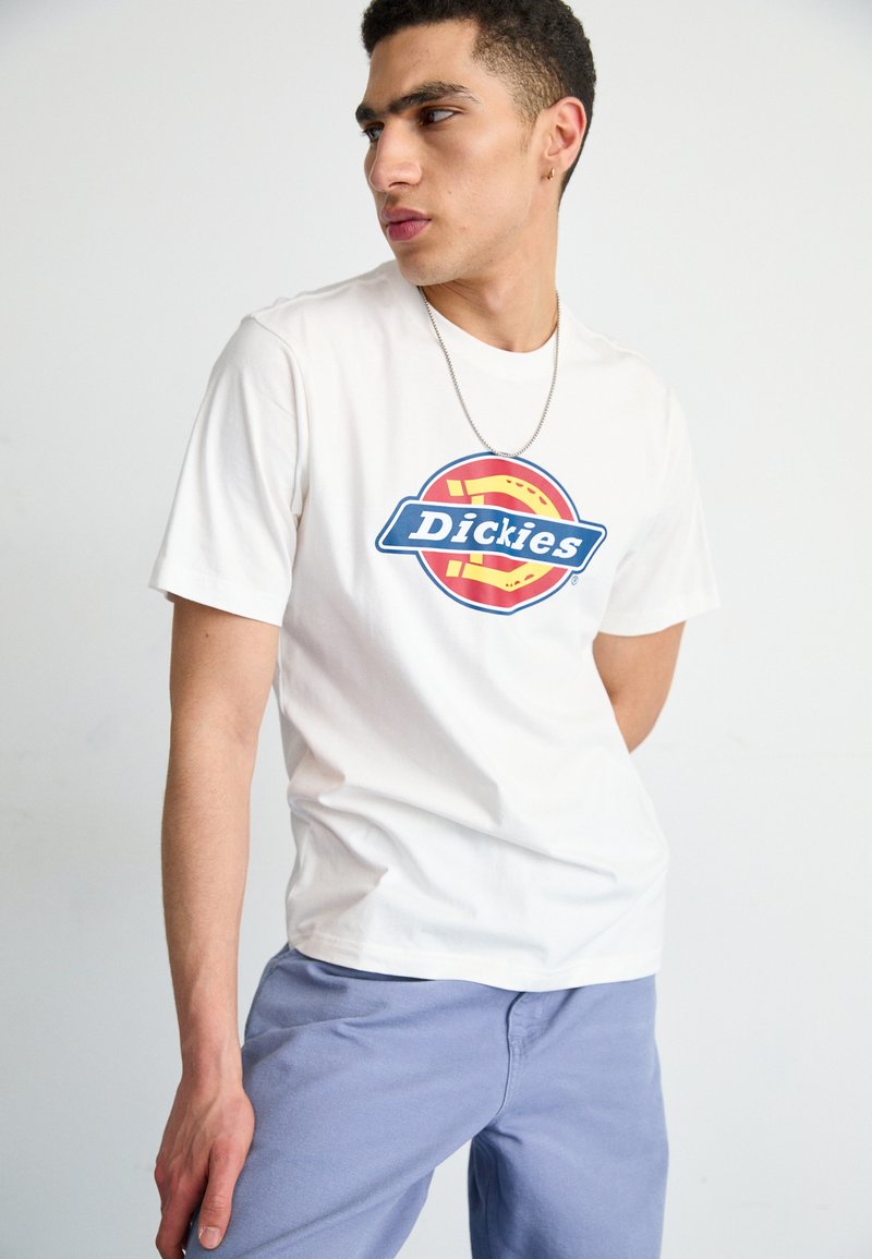 Weißes Baumwoll-T-Shirt mit einem rundem blauen und roten Dickies-Logo auf der Vorderseite. Das Modell trägt lila Hosen und eine silberne Kettenhalskette.