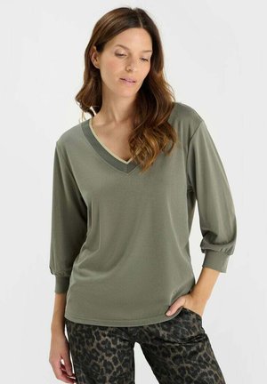 Woman wearing an olive green long-sleeve V-neck top and dark leopard print pants, with one hand in her pocket, looking downward.