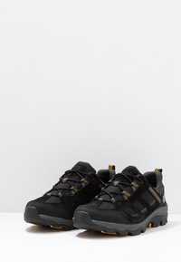 Scarpe da esterno nere con una combinazione di materiali in rete e suede, dettagli gialli, sistema di allacciatura e suola strutturata e robusta.