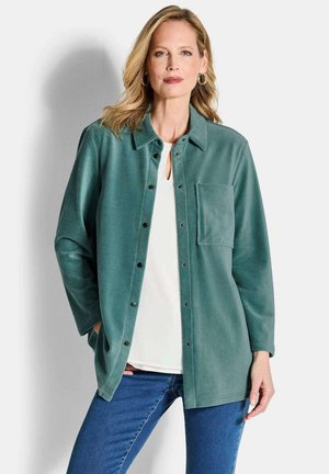 Teal corduroy shirtjas met een knoopsluiting aan de voorkant, een enkele borstzak en lange mouwen, gedragen over een wit topje en een blauwe spijkerbroek.