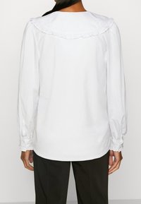 Blusa bianca a maniche lunghe con colletto arricciato e maniche arricciate. Realizzata in tessuto leggero, presenta una texture liscia e una vestibilità rilassata.