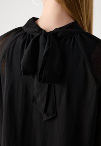 Svart texturerad blus med genomskinliga ärmar och en knuten rosettkrage som bärs av en person med långt rakt hår synligt bakifrån.