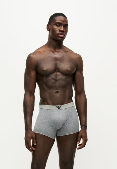 Homme musclé debout sur fond blanc, portant un boxer gris avec une ceinture de marque, regardant légèrement sur le côté.
